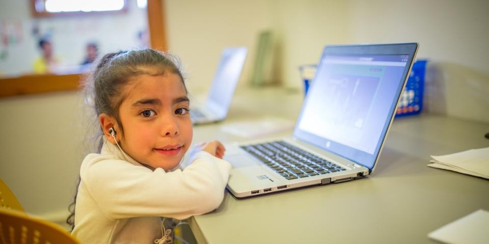 A young girl smiles at the camera while working on a laptop at a desk.