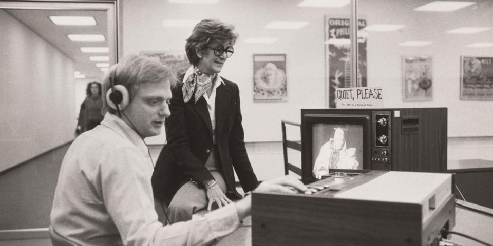  A black and white photo from 1981 of a man listening to audio through headphones, and a woman smiling looking at a television set, upon which a sign says: Quiet Please.