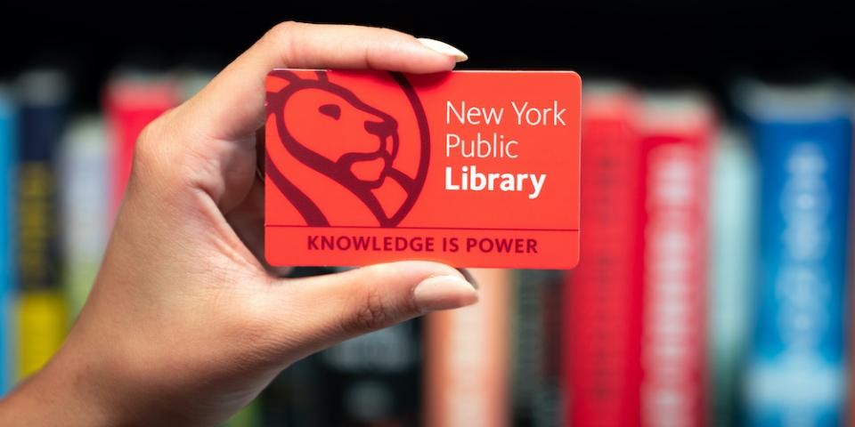 In front of a bookshelf, a hand holds an NYPL card that with a lion logo and text that reads: New York Public Library, Knowledge Is Power. 