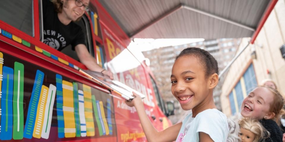 Several children smile and receive books from the NYPL Bookmobile.