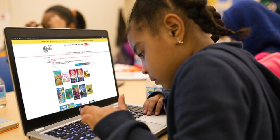 A young girl types on a laptop with an NYPL browsing window displayed on the screen.