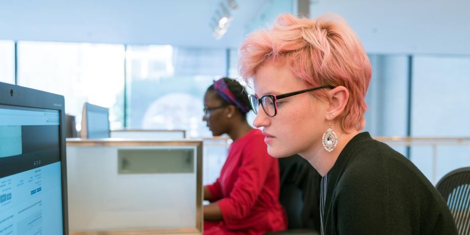 A woman with pink hair sits a computer, another woman sits at another computer in the background.