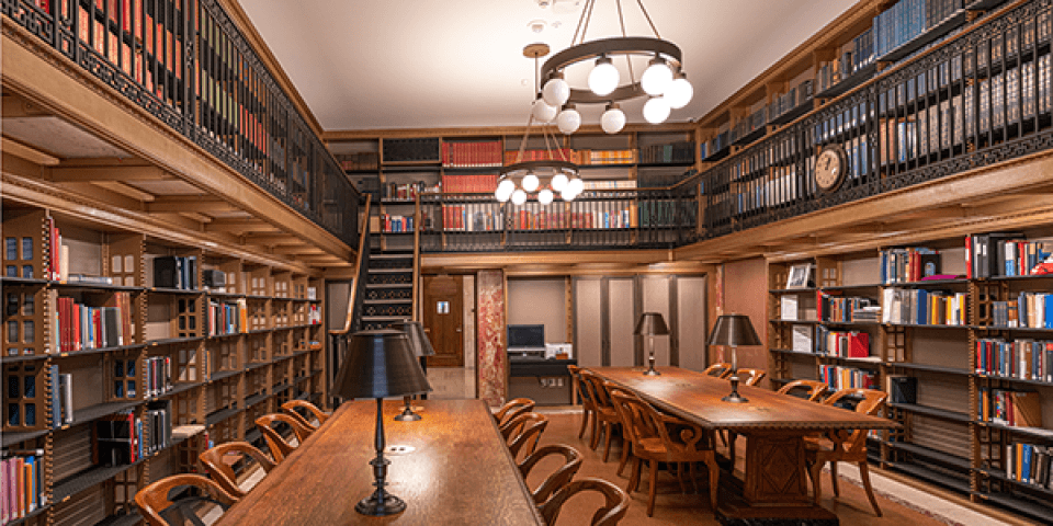 A room lined with two floors of bookshelves has long wooden desks with lamps and a printing station.