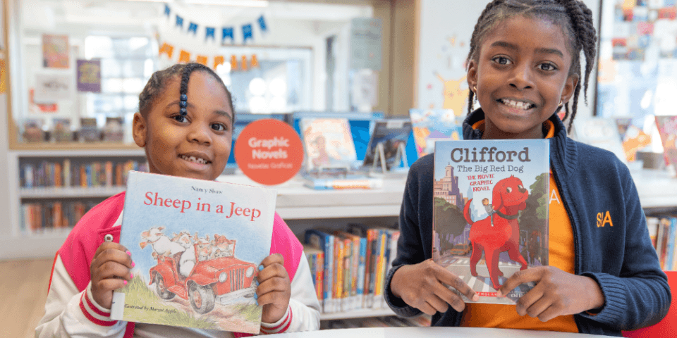 Two girls smiling in a classroom setting as they both hold up two books in their hands. 