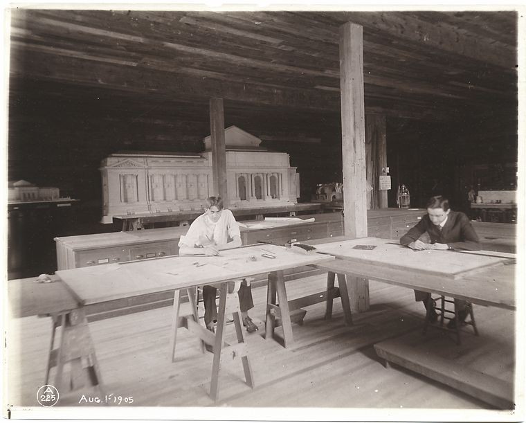 Two men sit in an open workshop with model of NYPL in background.