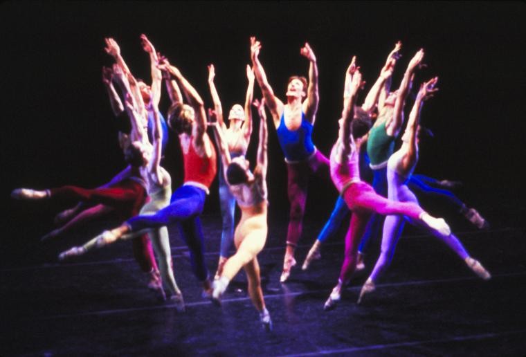 group of dancers on stage wearing colorful leotards stretching their arms up, one foot back, as they lean into the center