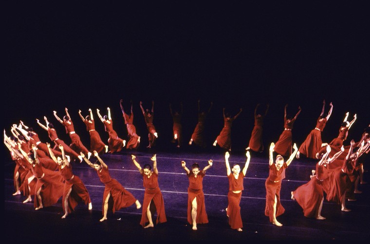 a circle of women dancers, arms outstretched, on stage all dressed in red