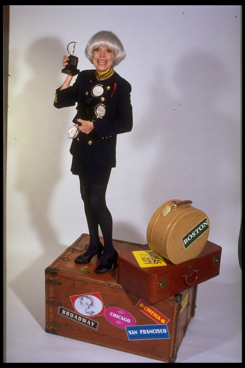 Carol Channing standing on a steamer trunk holding 4 TONY awards.