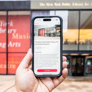 Hand holding a phone displaying the LPA Bloomberg Connects guide, with the library's facade in the background.