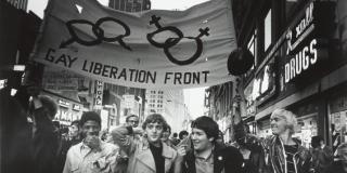 A crowd of people are shown marching down a street lined with storefronts and tall buildings. The five men in the foreground walk in line with each other, the two men on the left and right-most side hold up a large banner with the words "Gay Liberation Front", two interlocked mars symbols and two interlocked venus symbols printed on it 