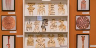 Several Large wooden combs sit arranged in a three row display case. Surrounding the case are photographs showing carved wooden combs and etched disks