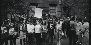 A large group of people, including Marcia P. Johnson, stand arm in arm in a black and white photo, some hold the Black Power fist in the air, protest signs for Gay Liberation can be seen in the background