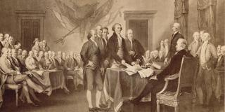 A colonial conference hall is shown. A smaller group of men, with Thomas Jefferson at center, gather around the Declaration of Independence, books and documents piled on a table in the foreground,  a larger group of men sit around the side of the room in the background. 