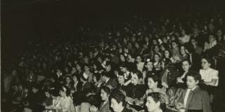 A large crowd of people is shown sitting in a large darkened theater