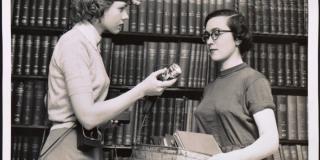 Black and white photograph of a woman wearing a headband light offering another woman a similar device.