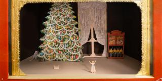 A photo of a set model with red fabric sides. The proscenium is made out of gold lace with pink, blue, and yellow jewels across the top. A window and bookcase is is the back right corner. A decorated Christmas tree is on the left with a mouse peaking out from behind it. A doll bed in front of the tree. A girl is standing in the front center, looking at the Christmas tree with her arms raised in the air.
