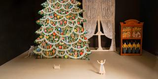 A close up photo of a set model. A window and bookcase is is the back right corner. A decorated Christmas tree is on the left with a mouse peaking out from behind it. A doll bed in front of the tree. A girl is standing in the front center, looking at the Christmas tree with her arms raised in the air.