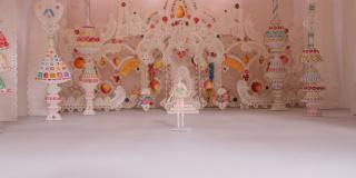 A close up photo of a set model. The interior of a castle with a throne is made out of white lace covered in colorful candy. A ballerina in a green tutu is standing the center.