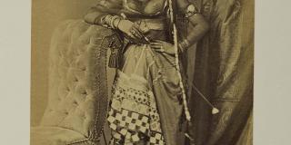 A sepia photograph of a young woman dressed in Indian garb. Her left hand rests on her hip and her right elbow rests atop a plush chair with a high back