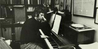 A black and white archival image of Stephen Sondheim sitting a piano in his office.