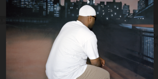 Man in white t-shirt with khaki pants and mustard colored workboots kneels in front of a painted canvans backdrop of a city skyline