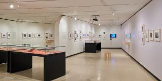 Photograph of a room in the exhibition with a curved wall dividing the room, three table cases, and framed photographs on the wall.