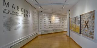 Exhibition entry way with yellow/brownish flooring and hanging artwork on the right wall and black text on a white wall with a title that reads Marking Time: Art in the Age of Mass Incarceration
