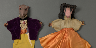 Two hand puppets with painted faces and hands, a mouse wearing a shirt and velvet coat and a cockroach wearing a veil, blouse, and dress.