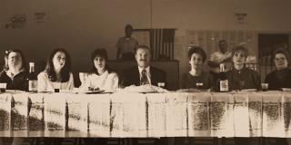 Photograph of seven people sit at a table facing the viewer. 