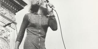 Photograph of Sylvia Rivera holding a microphone, speaking in front of Washington Square arch