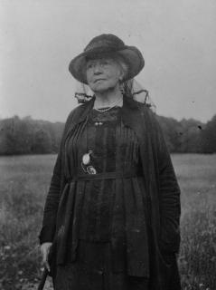 An elderly Lady Gregory standing alone in a field wearing all black 