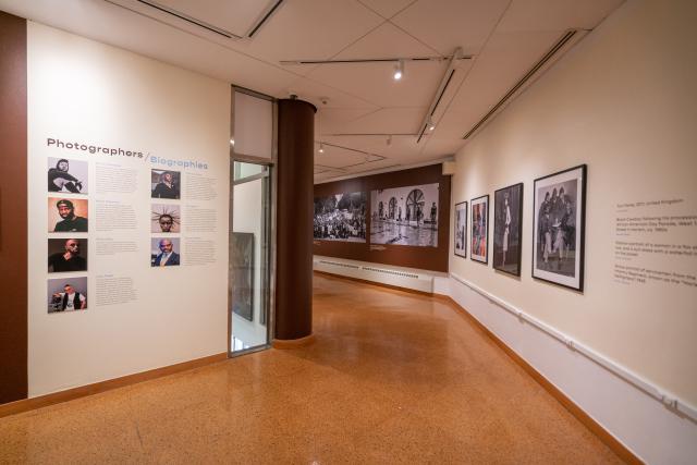 Wide angle image of a gallery. On the left are seven images and bios of photographers on a cream background, and on the right are framed photographs hung on a cream wall.