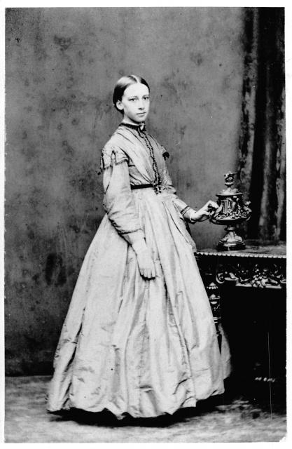 black and white photo of young Lady Gregory standing by desk wearing a dress