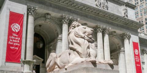Marble lion statue in front of the Stephen A. Schwarzman Building, which has two red banners.