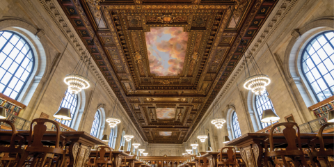 Perspective view of the Rose Main Reading Room, featuring an ornate ceiling, large arched windows, and wooden tables and chairs. 