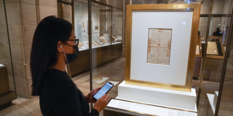 A woman in glasses and a black face mask holds her phone up while looking into a display case with Washington's farewell address. 