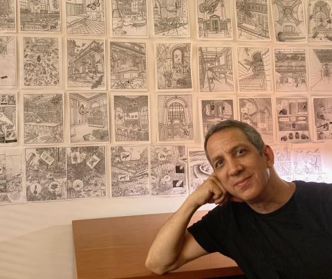 photo portrait of Peter Kuper sitting in front of a wall where his drawings hang