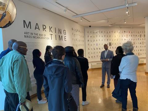 Group of diverse people on a tour of an exhibition with black text on a white wall that reads Marking Time: Art In the Age of Mass Incarceration