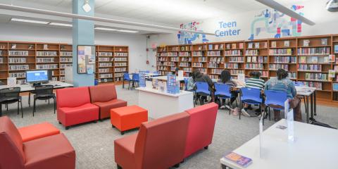 Baychester Library Teen Center, featuring red and orange lounge chairs, computer tables, work tables, and shelves of books lining the walls.